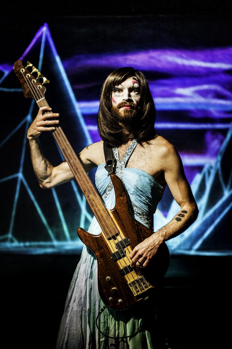 Aaron Collier_The Princess Show. A man in a dress, with long hair, a beard and a moustache, holding a guitar. There are laser like neon lights behind him.
