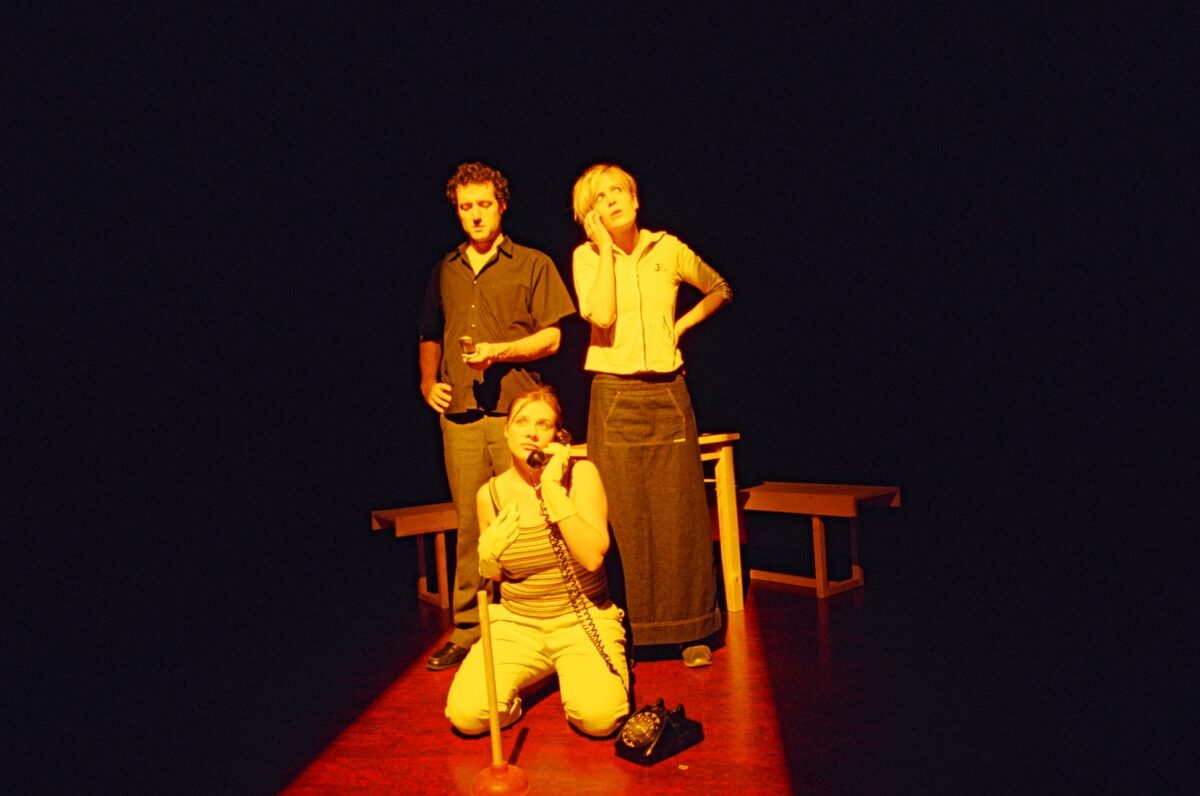Men of the World. Photo of three people standing on a stage with an orange spotlight shining on them. Two people are standing and the person in the middle in sitting on their knees and is using an old style phone.