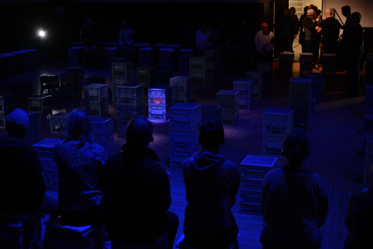 A dram image of a stage with milk crates staked everywhere. There's a small group of people at the top of the image with a faint light.