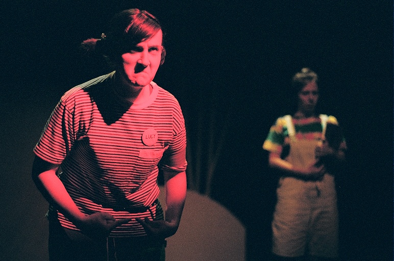 Wedgie 2. The photo shows two people on stage. A red spotlight is shining on the person on the left. They're wearing a striped T-shirt and a button that says 'Lucy'. They have their hair in pigtails. and are wearing glasses with a miserable expression. The person on the right is further back and blurry, they're wearing white overalls and a striped t-short.