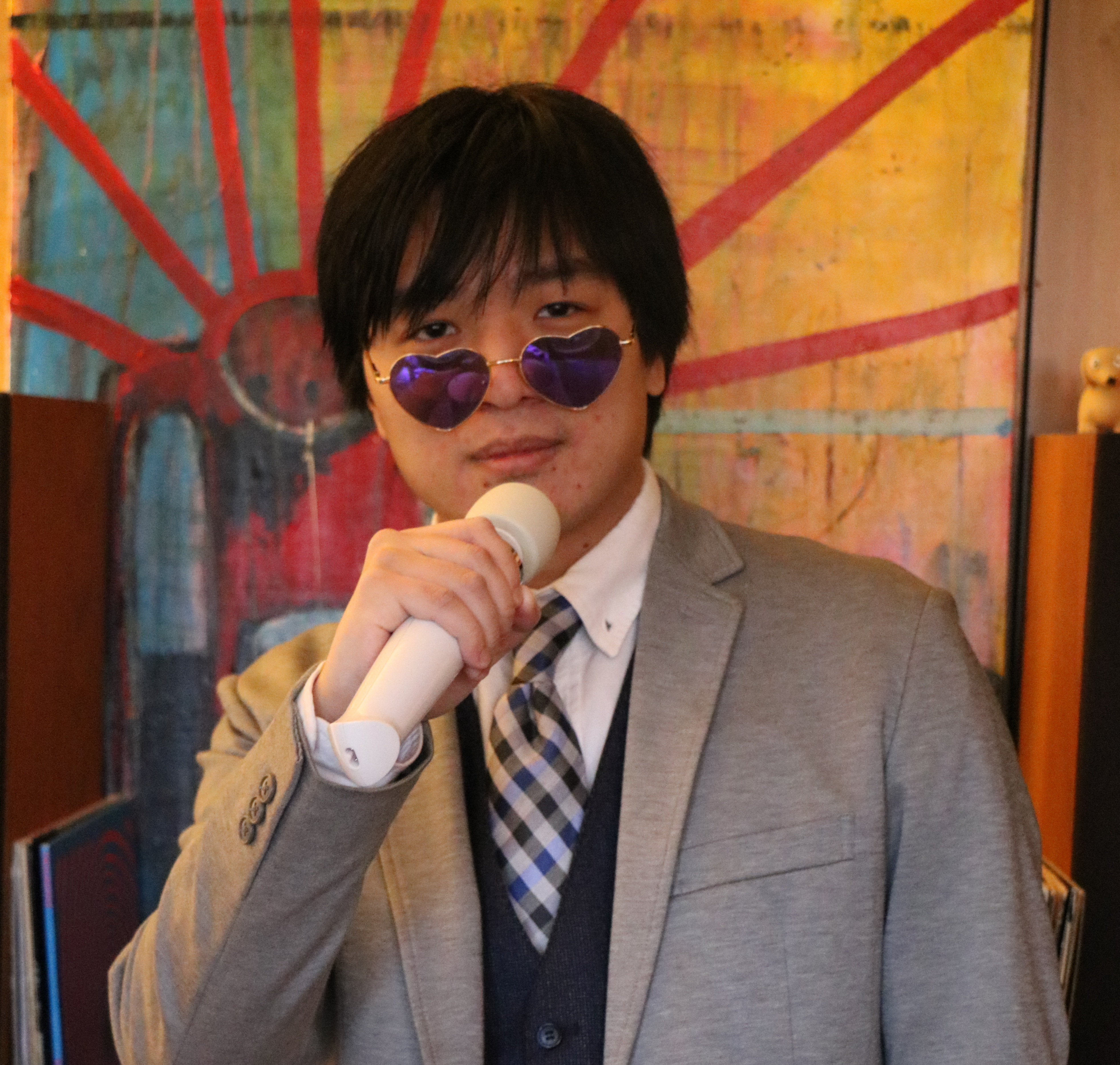 An asian man speaks into a microphone with purple heart shaped glasses on