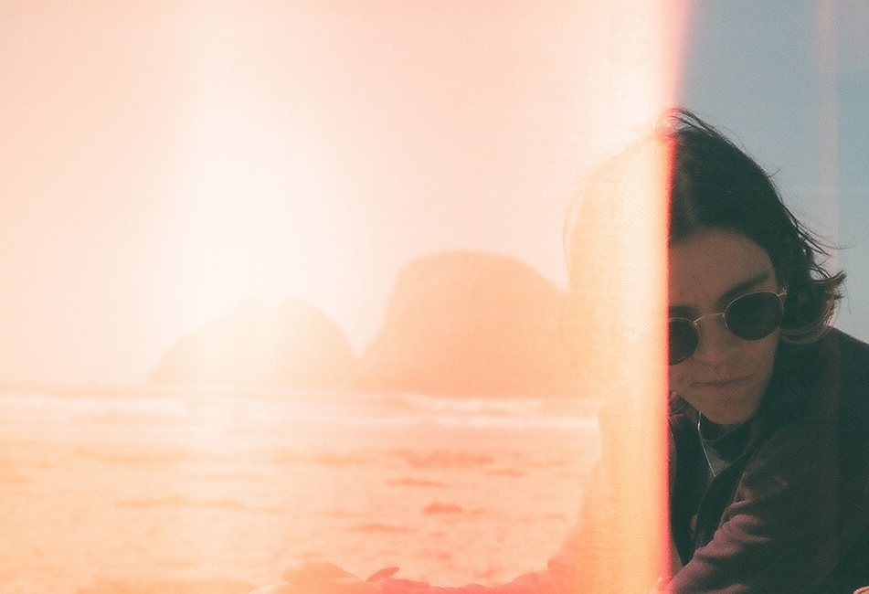 Photo is a white person with sunglasses on, looking down. The beach is out of focus behind them.