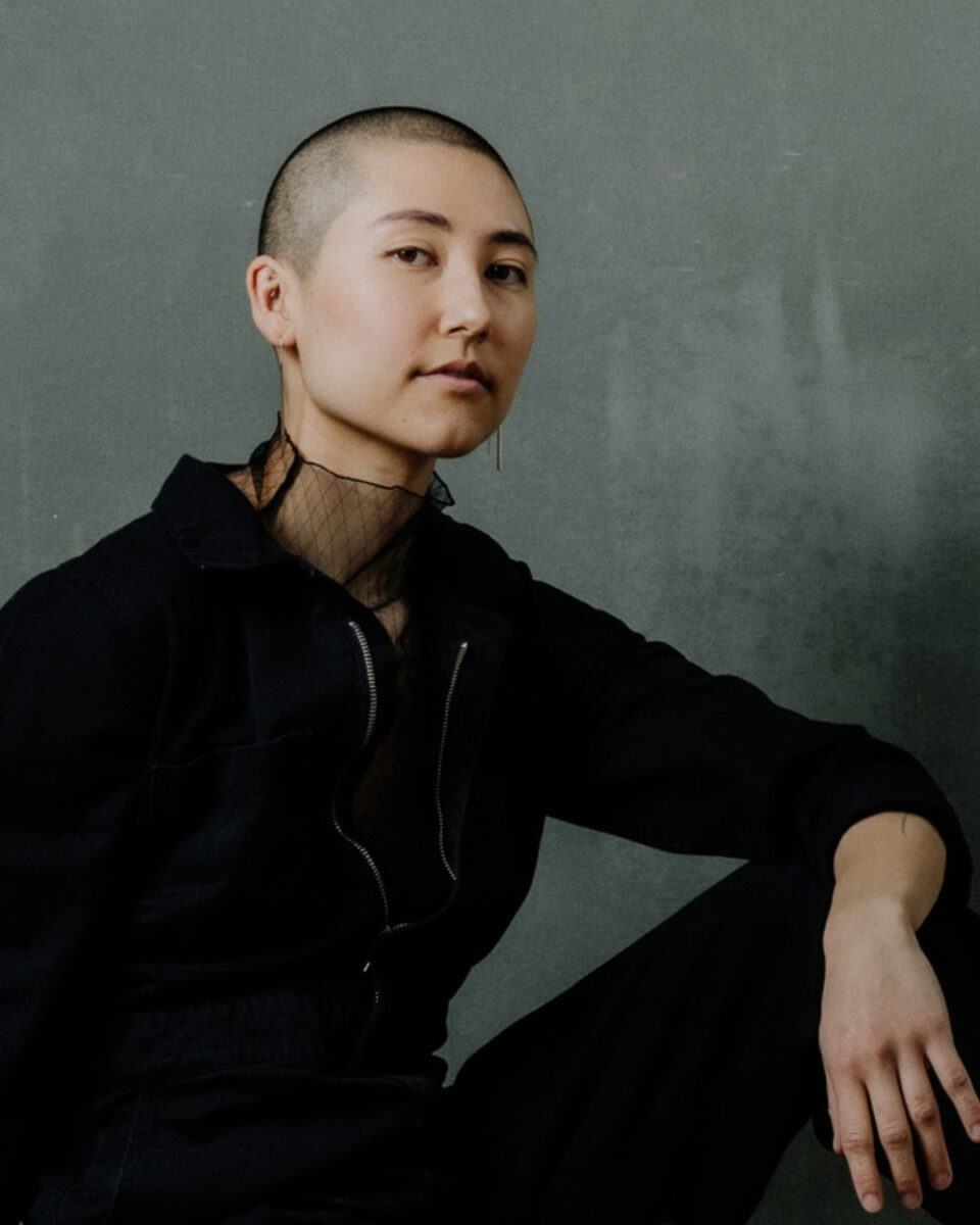 Headshot of a mixed-race person of Japanese and white European ancestry, with a shaved head wearing a black jumpsuit, leaning their left arm on their knee and looking into the camera.