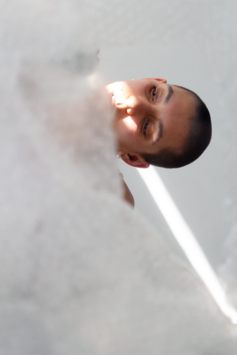 Headshot of a non-binary person with short shaved dark brown hair, framed by sheets of bubble wrap. Their head is tilted sideways, mid-motion. A thin streak of sunlight diagonally crosses their face.